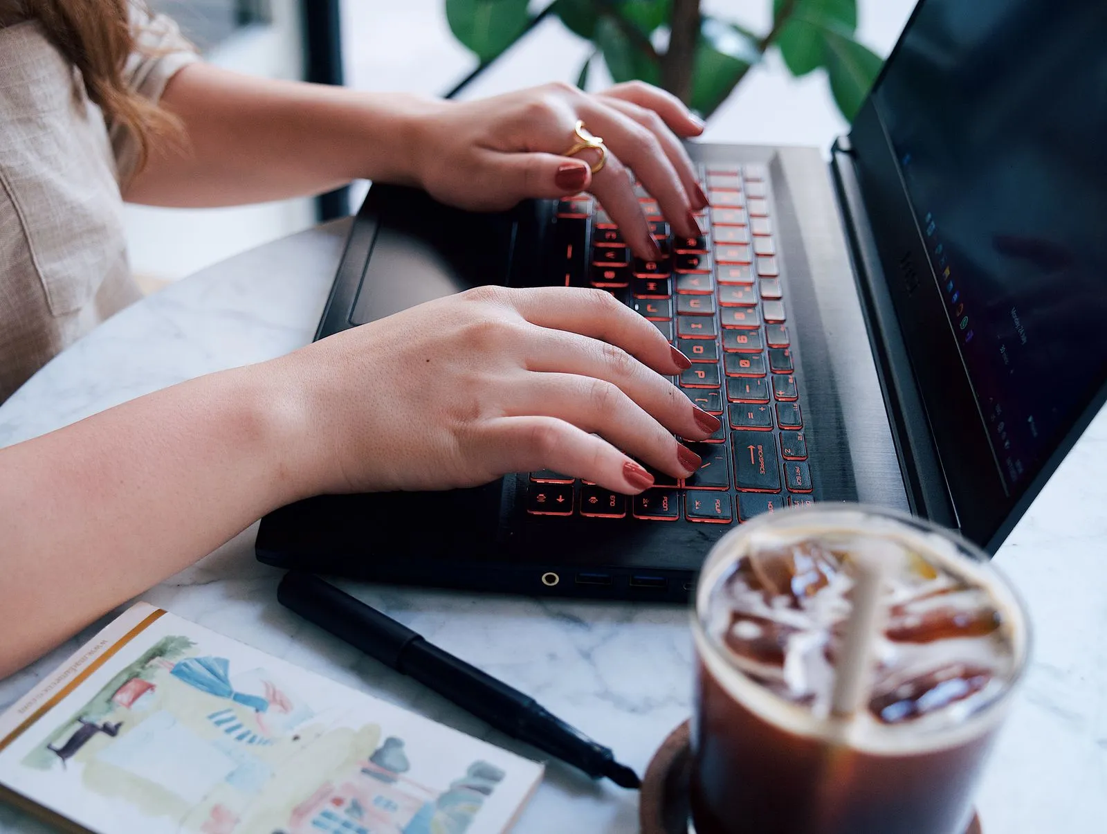 A young woman typing on a laptop at a café table with coffee and a notebook nearby.